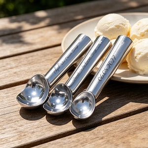 Könnte beinhalten: Drei polierte, silberfarbene Eisportionierer aus Metall mit zylindrischen Griffen. Der erste Griff ist mit 'Chyrl's Ice Cream Scoop', der zweite mit 'Pascale's Ice Cream Scoop' und der dritte mit dem Datum '06.08.2027' in Schreibschrift graviert.