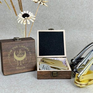 Könnte beinhalten: Ein Holzbox-Set mit einem goldfarbenen Wachssiegelstempel und einem passenden goldfarbenen Siegel. Die Box ist mit dem Text "FROM THE LIBRARY OF" und dem Namen "Elena" graviert. Zusätzliche Wachssiegelstempel sind sichtbar.