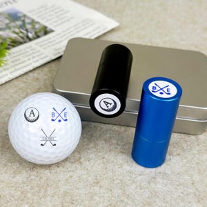 Könnte beinhalten: Ein weißer Golfball mit dem Buchstaben "A", gekreuzten Golfschlägern und dem Namen "LAUREL" darauf. Ein schwarzer und ein blauer zylindrischer Golfballstempel sind ebenfalls im Bild. Die Stempel haben den Buchstaben "A" und gekreuzte Golfschläger aufgedruckt.