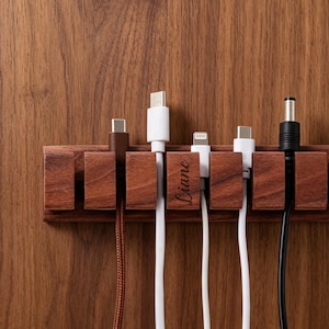 Op de afbeelding: Een kabelorganizer van donker hout met zes verticale sleuven voor diverse oplaadkabels. Eén houten segment is gegraveerd met de naam Liane in cursief schrift. De kabels bestaan uit een bruin gevlochten snoer, witte snoeren en een zwart snoer.
