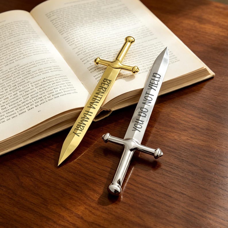 May include: Two sword-shaped metal bookmarks. One is gold-toned with the text "BERNINA HAMBLY" engraved on the blade. The other is silver-toned with the text "YOU DO NOT YIELD" engraved on the blade. Both feature a crossguard and pommel.