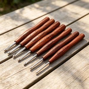 Peut inclure: Un ensemble de six crochets avec des pointes en métal poli et des manches en bois brun à grain. Chaque manche est gravé d'un nom unique et d'une mesure de taille, dont 2.0mm et 3.0mm, disposés en diagonale sur une surface en bois clair.