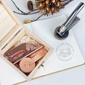 May include: A wooden box with a copper-colored letter opener and a round paper cutter. The letter opener has a floral design. A silver and black book embosser sits on a white notebook with a library stamp.
