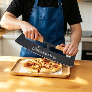 Könnte beinhalten: Ein schwarzer Pizzaschneider mit zwei hellen Holzgriffen und dem weißen Schriftzug "Christian Piscos" auf der Klinge. Der Schneider ist über einer belegten Pizza positioniert, die auf einem rechteckigen Serviertablett aus Bambus liegt.