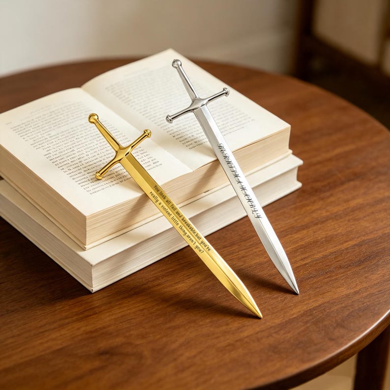 May include: Two metal sword-shaped bookmarks resting on an open book. One bookmark is gold-toned with the engraved text: You look all frail and breakable, but you're really a violent little thing, aren't you? The other is silver-toned with engraved text.