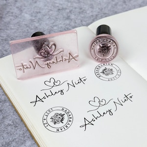 Peut inclure: Tampons transparents avec le nom "Ashley Nieto" en écriture cursive et un motif de cœur. Un tampon rond avec un motif floral et le texte "BAHAR & ARIAN" est également visible. Les tampons sont sur une surface blanche.