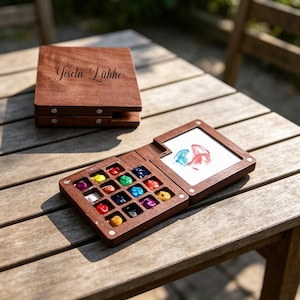Peut inclure: Un coffret de peinture aquarelle en bois avec un couvercle à charnière portant le nom gravé Gisela Lübke. Le boîtier ouvert présente seize compartiments carrés remplis de peintures multicolores et une zone de mélange avec des traces.