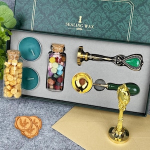 Könnte beinhalten: Ein Siegelwachs-Set in einer grün-goldenen Box. Das Set enthält teal Kerzen, Wachsperlen in einem Glas, einen Wachsstempel mit einem grünen Stein, einen Wachsschmelzlöffel und einen goldenen Stempelständer. Auf dem Deckel der Box steht "SEALING WAX".