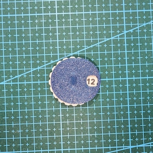 Peut inclure: Un petit objet rond bleu avec un bord festonné et le chiffre 12 imprimé dessus. L'objet a une découpe rectangulaire au centre et est posé sur un fond de grille verte.