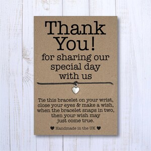 以下が含まれることがあります： 「Thank You! for sharing our special day with us」と書かれた茶色のカード。カードには、銀色のハートのチャームが付いた黒い紐のブレスレットが付けられています。カードには、「Tie this bracelet on your wrist, close your eyes & make a wish, when the bracelet snaps in two, then your wish may just come true. Handmade in the UK.」という文章も書かれています。