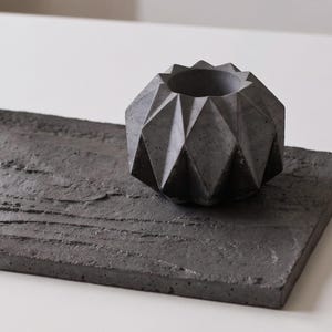 May include: A dark gray rectangular concrete tray with a textured, uneven surface paired with a matching dark gray concrete planter featuring a geometric, faceted design with multiple triangular planes and a circular opening at the top.