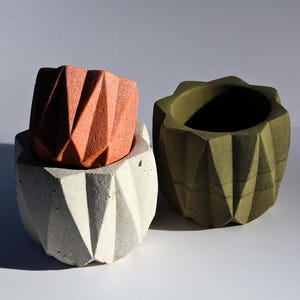 May include: Three geometric concrete planters in shades of orange, white, and olive green. The planters have a faceted design, with the orange planter stacked on top of the white one. The olive green planter stands alone.