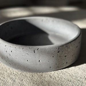 May include: A gray concrete bowl with a thick, rounded rim. The bowl has a rough, textured surface with small holes. The bowl is sitting on a concrete surface. The bowl is likely used for home decor.