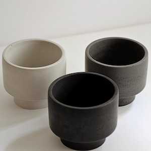 May include: Three cylindrical planters with a stepped base design. One planter is light beige, one is medium gray, and one is dark charcoal gray. Each vessel features a smooth, matte finish and a wide, open top, arranged in a cluster formation.