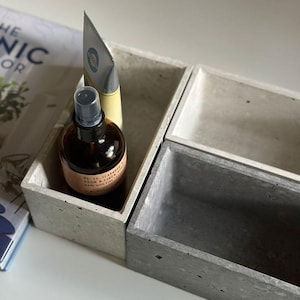May include: Two concrete rectangular trays with rounded edges. The trays are in different shades of gray. One tray contains a clear glass bottle with a brown liquid and a silver tube with a green label. The bottle has a black label with white text that reads "No. 06 - TEAKWOOD ROOM & LINEN SPRAY MADE WITH ESSENTIAL OILS".