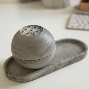 May include: A gray concrete oval tray with a smaller gray concrete sphere with holes on top. The sphere is sitting in the center of the tray.