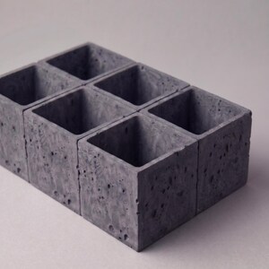 May include: A set of six connected square containers made of dark gray concrete. Each container features a porous, textured surface with visible air bubbles and a hollow interior, arranged in a two-by-three grid formation with sharp edges.