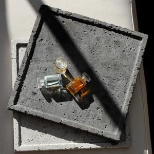May include: Two square concrete trays, one dark gray and one light gray, stacked together. Three small glass bottles containing clear, light green, and amber liquids are placed on the top dark gray tray, casting sharp shadows.