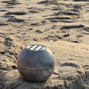 May include: A gray concrete sphere with a circular opening and small holes around the opening. The sphere is sitting on a sandy surface.