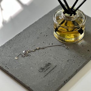 May include: A gray concrete tray with a silver chain necklace with star charms. The tray has the text "Olesia in design" and a website address. A glass bottle with black sticks is on the tray.