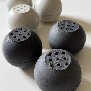 May include: Six spherical, decorative objects. Three are light gray, and three are dark gray. Each has a textured surface and a circular top with several small holes. The objects are arranged on a white surface.