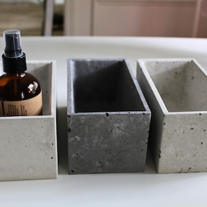May include: Three rectangular concrete containers in varying shades of gray. One container holds a brown glass spray bottle with a black top and a label. The other two containers are empty. The containers are arranged on a white surface.