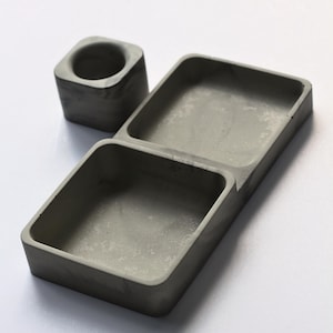 May include: A gray concrete desk organizer with two rectangular compartments and a small square candle holder. The organizer is made of a smooth, solid material and is designed to hold small items. The items are gray.