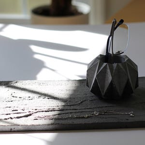 May include: A dark gray concrete desk organizer set. The set includes a geometric-shaped pen holder with eyeglasses and a rectangular tray with a silver necklace. The tray has a textured surface.