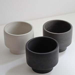 May include: Three modern, cylindrical planters in varying shades of gray. The planters have a rounded base and a wide opening, perfect for small plants or succulents. The minimalist design and neutral colors make them suitable for any decor.
