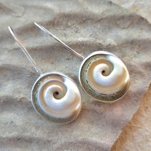May include: A pair of drop earrings featuring circular shiva eye shell centers with a natural white spiral pattern, surrounded by a textured olive-green border, set in polished silver-toned metal frames with long, slender silver-toned ear wires.