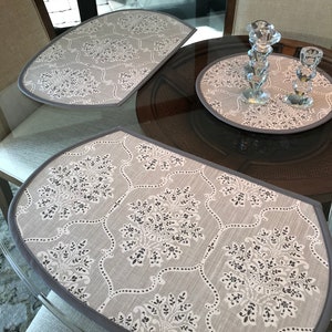 May include: Three gray placemats with a white floral pattern and a gray border are displayed on a glass table. Two clear crystal candle holders sit on a matching round placemat. The placemats are a decorative element for a dining table.