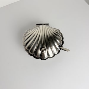 May include: A silver-toned metal condiment server shaped like a scallop shell with a hinged lid and a small matching spoon. The exterior features a ribbed texture radiating from the hinge, and a small spherical knob is attached to the front lid.