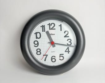 May include: A round, black wall clock with a white face and large black numerals. The clock has a red second hand and black hour and minute hands. The word "bino" is printed above the center of the clock face, and "quartz" is printed below.