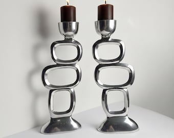 May include: Two silver-colored candle holders with a unique, geometric design. Each holder features a series of connected oval shapes, topped with a cup-like structure holding a dark brown candle. The candles are lit, casting a warm glow.