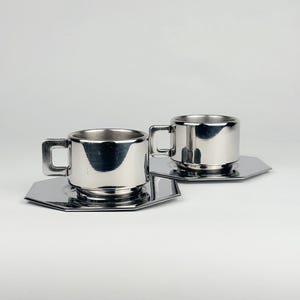 May include: Two stainless steel espresso cups with square handles sit on matching hexagonal saucers. The cups and saucers have a shiny, reflective surface. The set is designed for serving coffee or espresso.