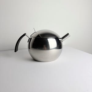 Könnte beinhalten: Eine kugelförmige Teekanne aus Edelstahl mit polierter, reflektierender Oberfläche. Sie verfügt über einen geschwungenen schwarzen Griff an der Seite, einen passenden gewölbten Deckel mit einem kleinen Metallknauf und einen kurzen, geraden Ausguss.