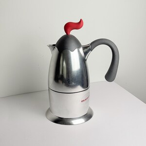 Puede incluir: Cafetera espresso de acero inoxidable con asa negra y parte superior roja en forma de llama. La cafetera tiene un acabado plateado y una parte superior negra. La marca "Bugatti" está impresa en rojo en el lateral.