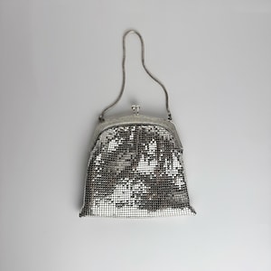 Pode incluir: Uma bolsa de mão em malha metálica prateada com uma alça de corrente flexível e um fecho de moldura articulada. O corpo da bolsa é construído a partir de pequenos quadrados metálicos interligados que criam uma superfície texturizada.