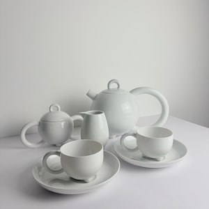 Könnte beinhalten: Ein weißes Keramik-Teeservice, bestehend aus einer Teekanne, einem Sahnekännchen, einer Zuckerdose und zwei Tassen mit Untertassen. Das Set hat ein minimalistisches Design mit abgerundeten Formen und glänzendem Finish. Die Tassen und die Zuckerdose haben kleine Füße.