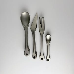 May include: A set of four matte silver-toned metal utensils arranged vertically, including a large spoon, a knife, a fork, and a small spoon. Each piece features a rounded handle with a circular hole at the base for hanging storage.