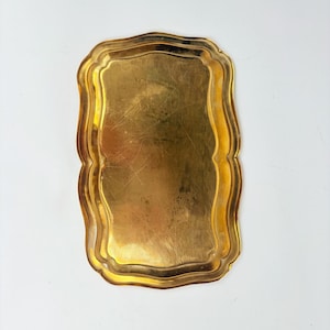 May include: A rectangular gold-colored tray with a scalloped edge. The tray has a slightly worn appearance, with visible scratches on its surface. The overall shape is a rectangle with gently curved sides, and the edges are adorned with a decorative, wavy pattern.