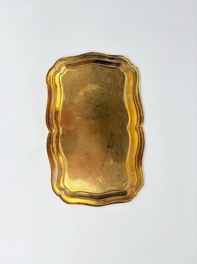 May include: A rectangular gold-colored tray with a scalloped edge. The tray has a slightly worn appearance, with visible scratches on its surface. The overall shape is a rectangle with gently curved sides, and the edges are adorned with a decorative, wavy pattern.