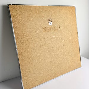 May include: A large, rectangular corkboard with a silver metal frame. The back of the board is visible, showing a metal hanger. The corkboard is set against a white background.