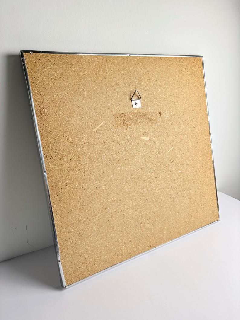 May include: A large, rectangular corkboard with a silver metal frame. The back of the board is visible, showing a metal hanger. The corkboard is set against a white background.
