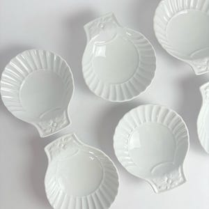 May include: Six white ceramic shell-shaped dishes are displayed. Each dish has a scalloped edge and a floral design on the handle. The dishes are arranged on a white surface, creating a clean and elegant aesthetic.