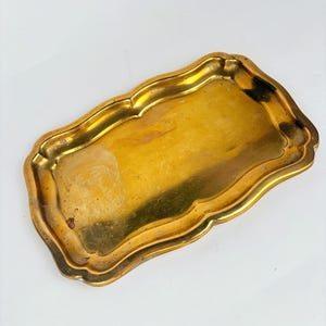 May include: A rectangular, antique-style brass tray with a scalloped edge. The tray has a tarnished, golden finish, showing signs of age and use. The tray is empty and sits on a white surface.