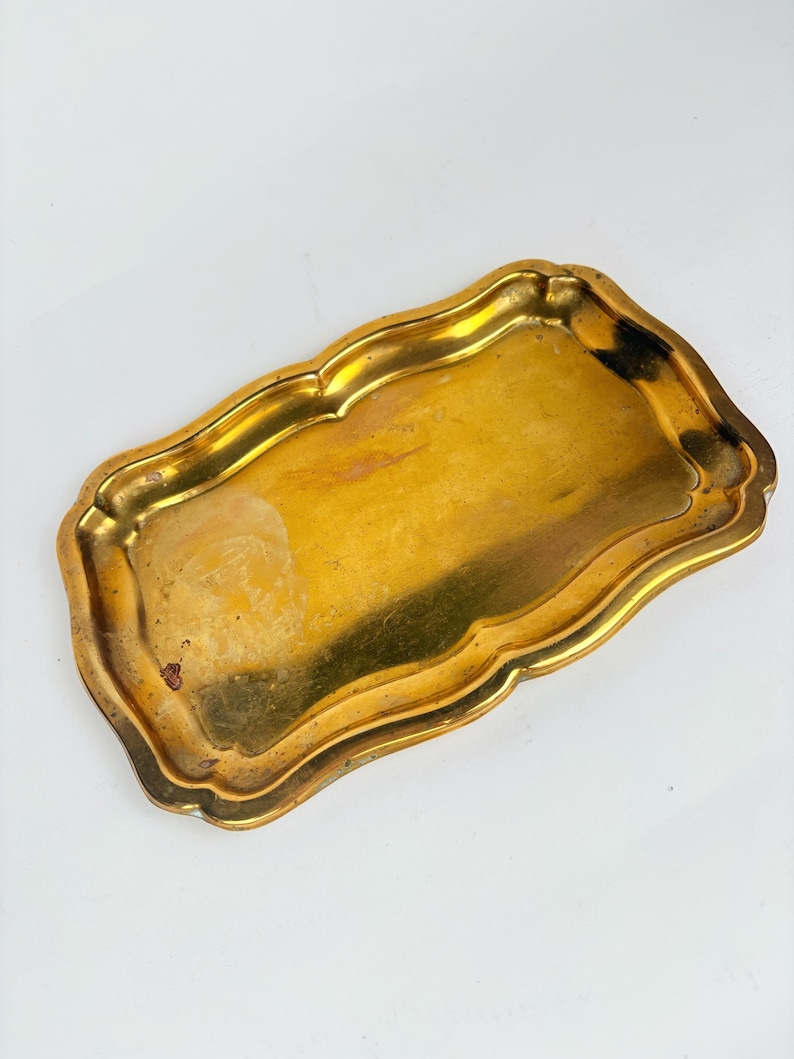 May include: A rectangular, antique-style brass tray with a scalloped edge. The tray has a tarnished, golden finish, showing signs of age and use. The tray is empty and sits on a white surface.