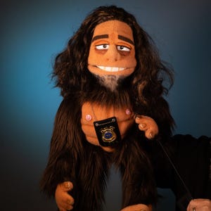 May include: A handmade puppet with brown fur, long dark hair, and a smiling face. The puppet has a badge around its neck and is holding a control stick. The text "HANDMADE BY @MYCUSTOMPUPPET" is displayed on a red banner.