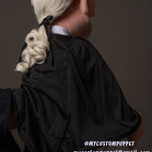 May include: A black puppet with a white wig and a black robe. The puppet is wearing glasses and has a black ribbon tied in its hair. The puppet is looking to the right. The text "@MYCUSTOMPUPPET mycustompuppet@gmail.com" is visible on the bottom right corner of the image.