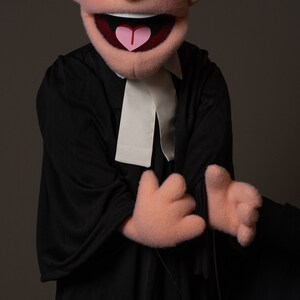 May include: A red-haired puppet wearing a black robe with a white collar. The puppet has a big smile and is holding its hands up in front of it. The puppet is being held by a hand with a ring on it. The text on the image reads "@MYCUSTOMPUPPET mycustompuppet@gmail.com".